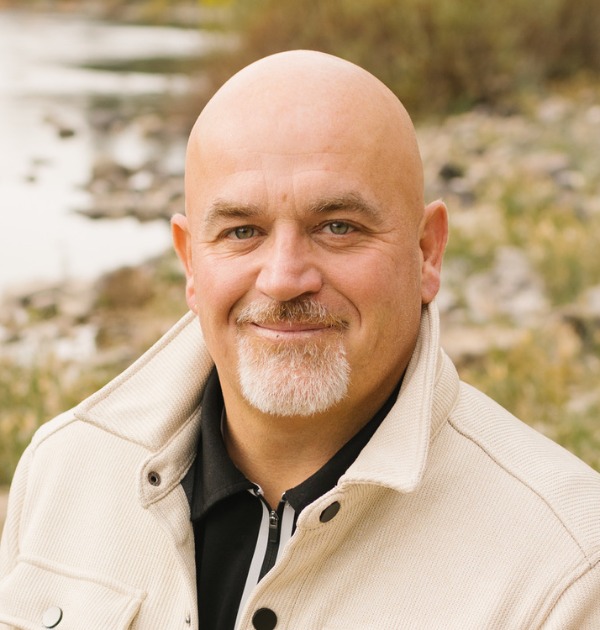 Mike Baker outdoors by the water in Coeur d'Alene, in a cream jacket, mid-smile.