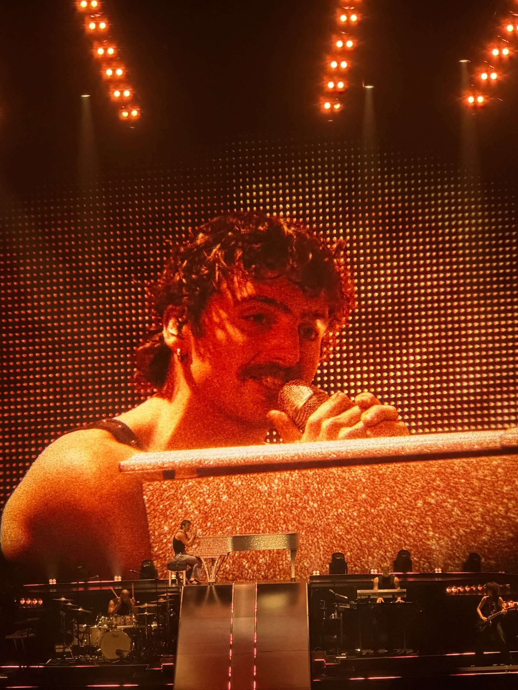 Benson Boone singing into a microphone on stage, shown large on the arena screen under orange lights