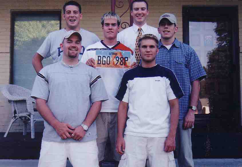 Six young men on a porch in the late 1990s, one holding up a Utah license plate that reads BGO 98P.