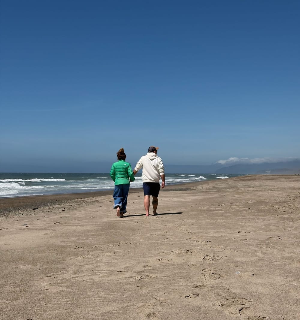 Mike Baker and his wife Kellie walking barefoot away down a wide, empty stretch of Pacific beach, holding hands, under a clear blue sky.