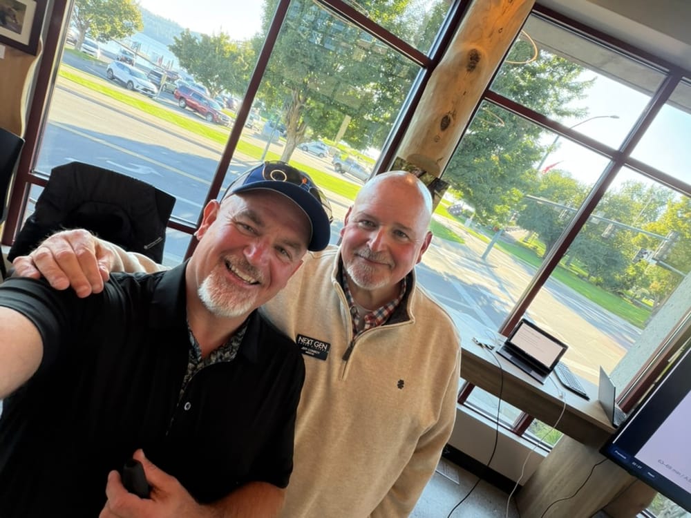 Mike Baker and Jeff Conroy smiling together in front of the windows before the NEXTGEN session, Jeff wearing his NEXT GEN name badge.