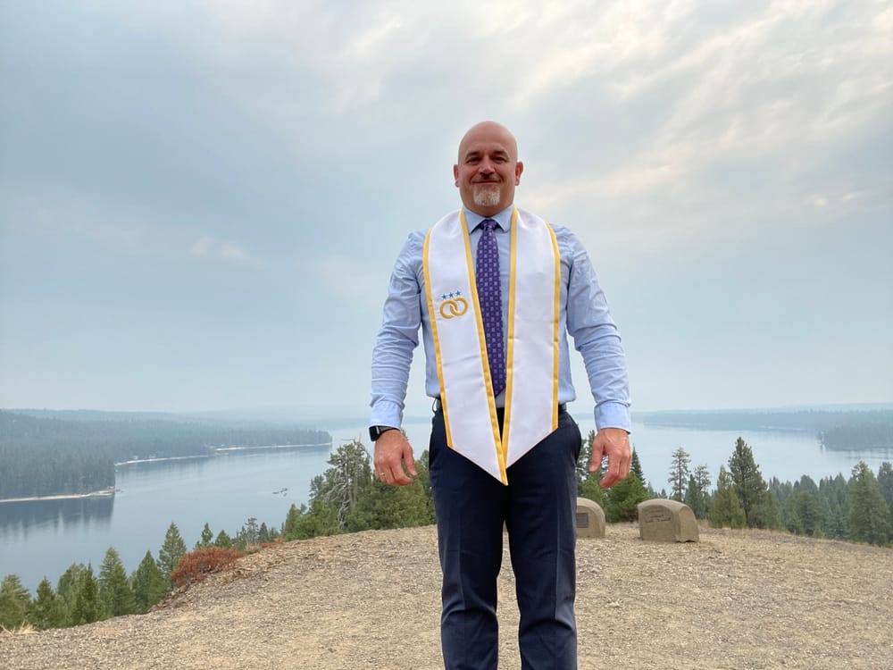 Mike Baker in a light blue shirt and tie wearing a white and gold American Marriage Ministries stole, standing on a hilltop above a North Idaho lake.