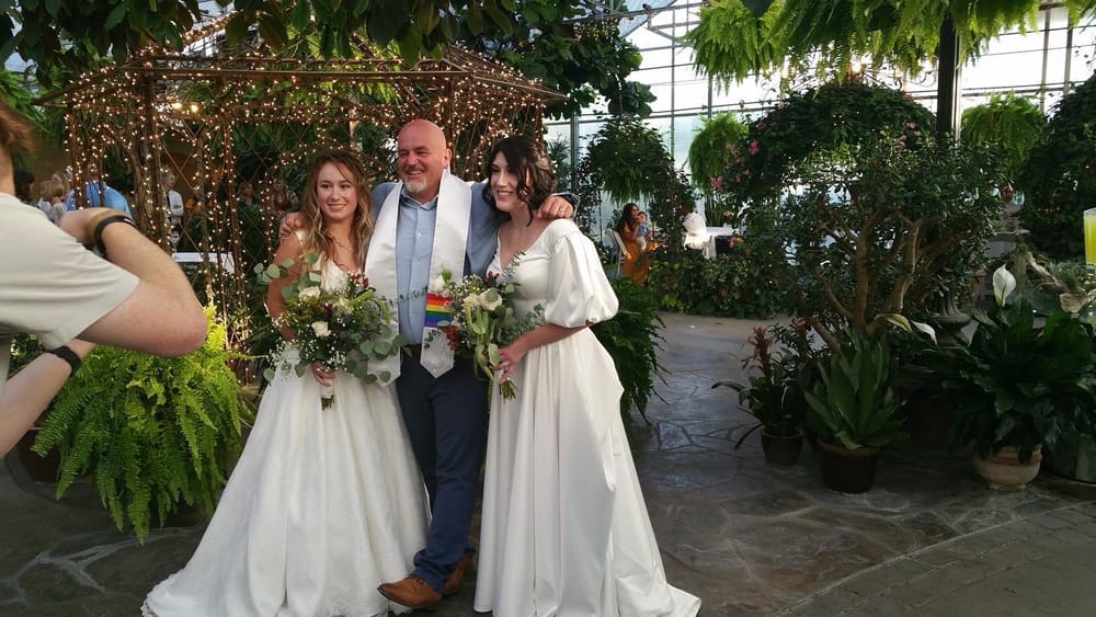 Mike Baker standing between two smiling brides in white dresses holding bouquets, wearing a rainbow officiant stole, inside a greenhouse strung with lights.