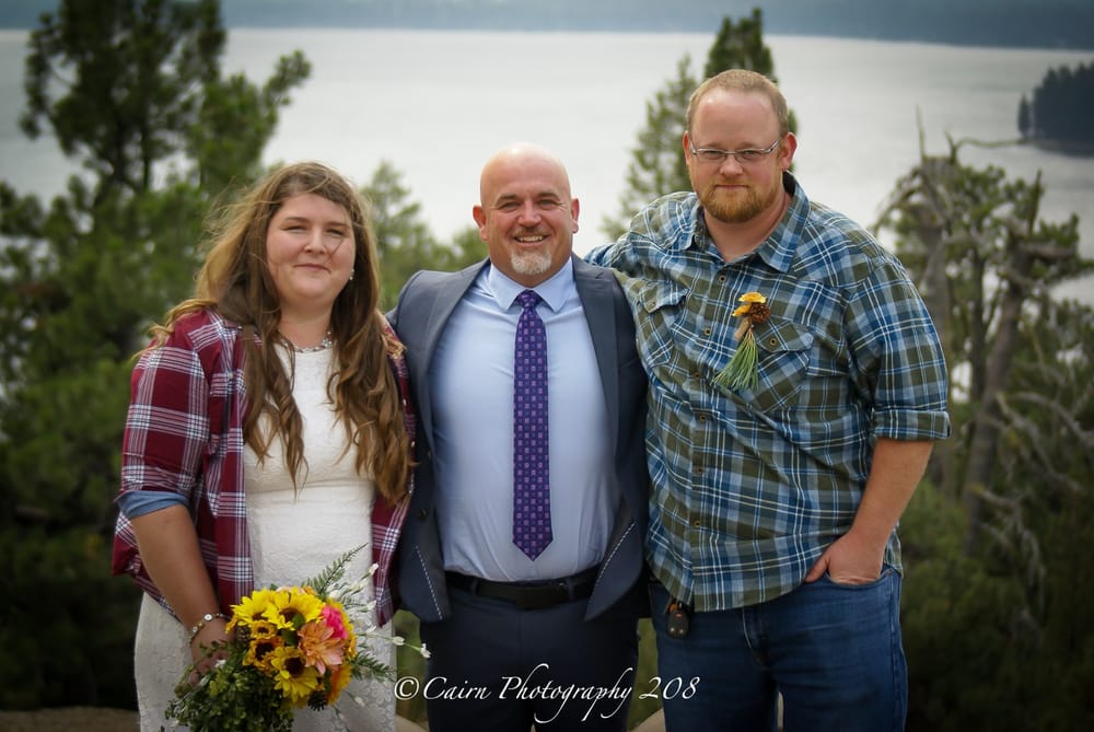 Mike Baker in a gray suit and purple tie between a newly married couple above a lake in McCall, Idaho, with a Cairn Photography 208 watermark.