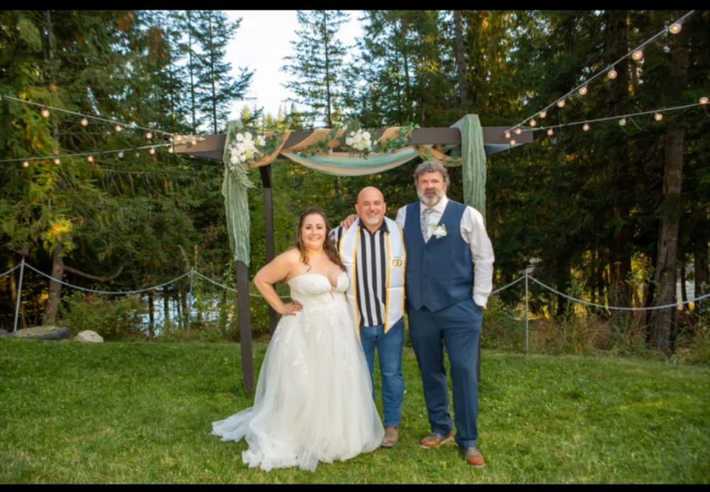 Mike Baker in a black and white striped referee shirt under an officiant stole, standing with a newly married couple beneath a wooden arch strung with lights in the woods.