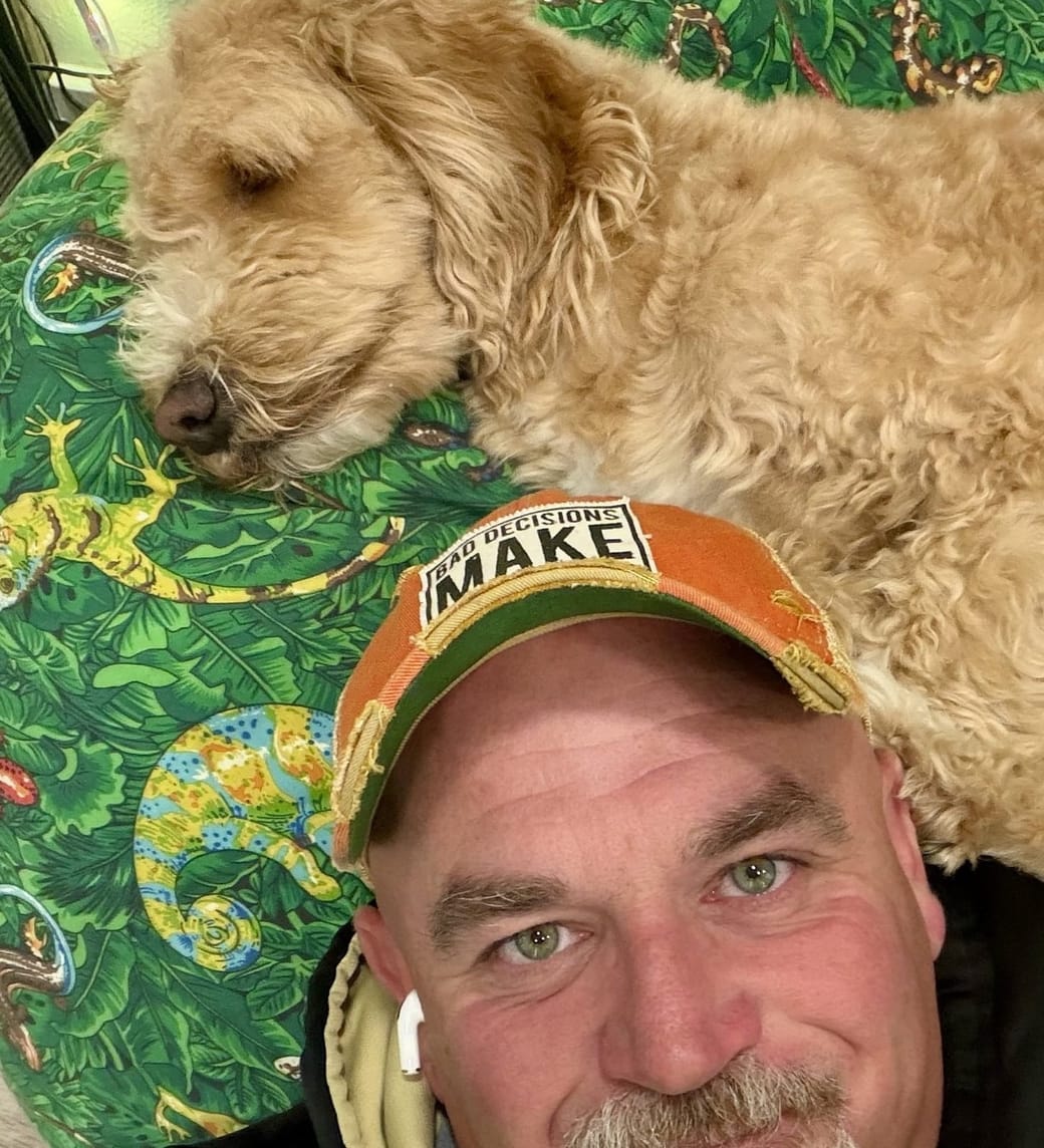 Mike Baker in a ball cap and hoodie, with Gus the dog asleep on the beanbag behind him