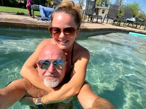 Kellie hugging Mike from behind in a swimming pool on a sunny day, both smiling in sunglasses.
