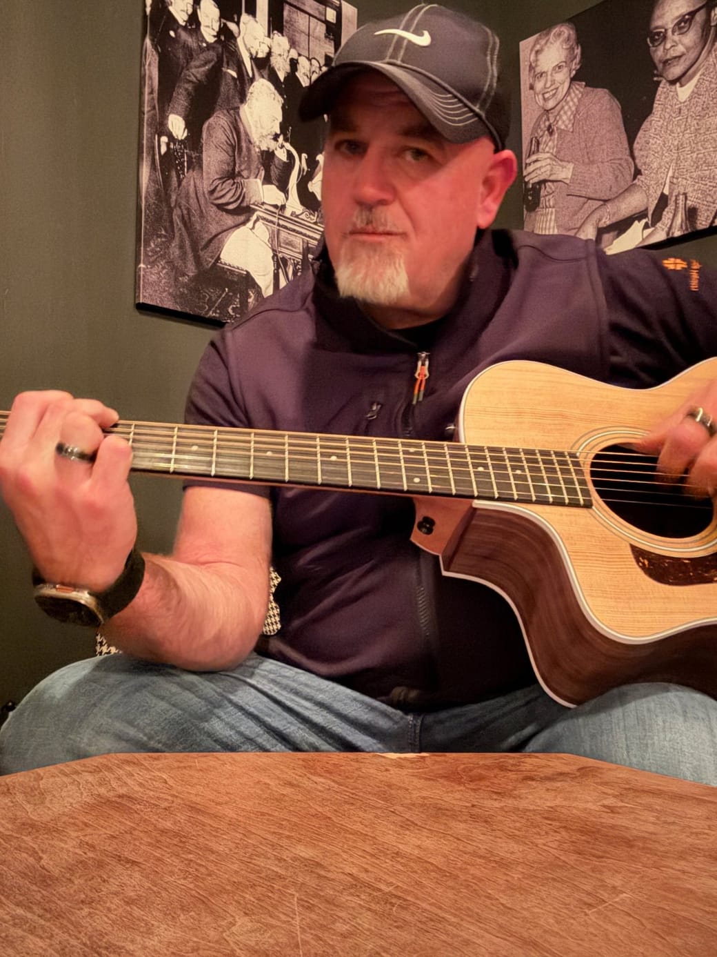 Mike Baker in a ball cap playing an acoustic guitar at a wooden table