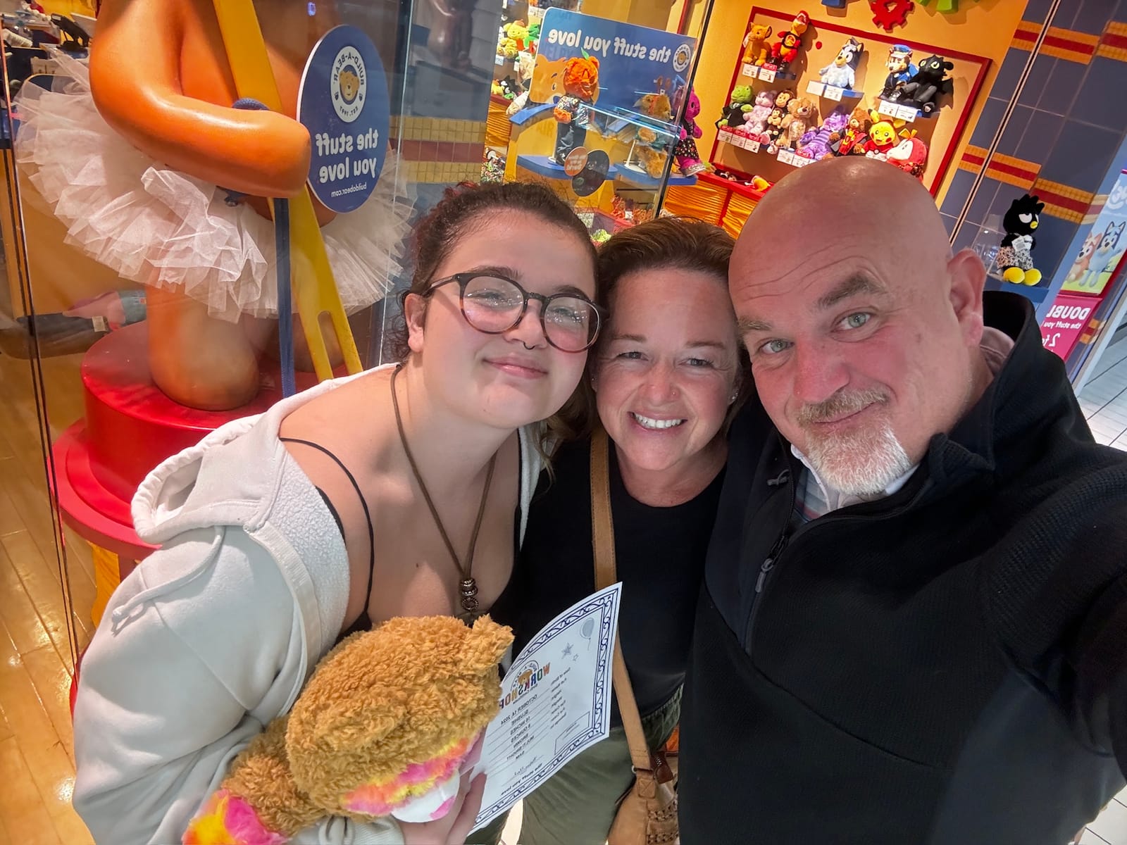Sammie, Kellie, and Mike Baker smiling together at Build-A-Bear, Sammie holding a new stuffed bear and its birth certificate.